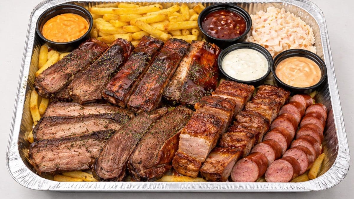 A BBQ smokehouse platter loaded with sliced brisket, ribs, pork belly, smoked sausage, fries, coleslaw and house sauces